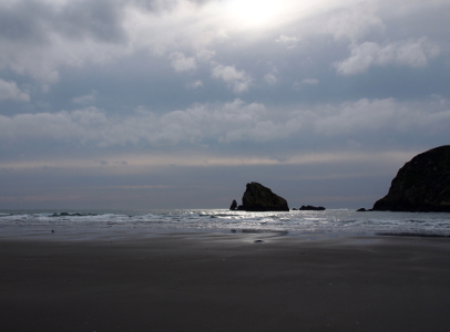 [A section of water just in front of one sea stack rock is lit while the area around it is darkened by clouds.]